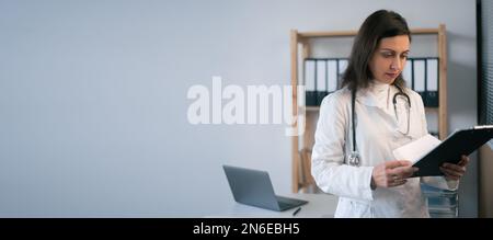 Services médicaux de qualité. Une femme médecin se focalisait dans une robe blanche pour lire des notes dans une planchette à pince alors qu'elle se trouvait dans son bureau d'hôpital. Copier l'espace. Bannière Banque D'Images