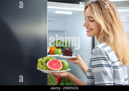 Concept de choix. Femme prenant des assiettes avec des fruits et des gâteaux du réfrigérateur dans la cuisine Banque D'Images