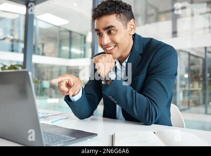 Jeune homme d'affaires de course mixte heureux pointant du doigt tout en travaillant sur un ordinateur portable seul au travail. Homme d'affaires hispanique souriant et lisant un Banque D'Images