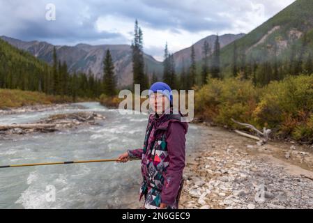 Une jeune pêcheur se tient sur les rives de la rivière alpine Shavla et attrape des poissons dans les montagnes de l'Altaï sous les nuages pluvieux près d'une forêt en Sibérie. Banque D'Images