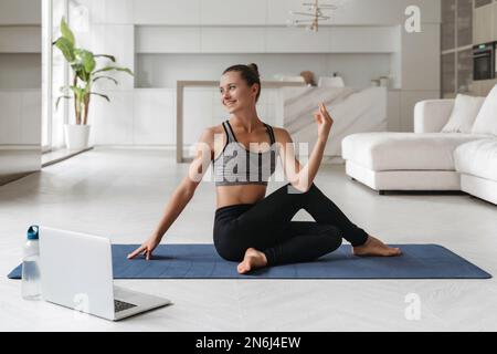 Les femmes font des exercices de torsion assis pour la santé et un corps en forme. Yoga à la maison, fitness, concept d'entraînement. Belle fille souriante regardant des tutoriels sur ordinateur portable, a cours de yoga en ligne à la maison dans sa chambre Banque D'Images