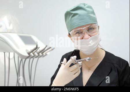 Femme médecin dans un cabinet dentaire en masque et lunettes capuchon vert pour contenir cheveux costume noir dans ses mains seringue carpienne et gants chirurgicaux hygiéniques mur blanc la dernière technologie croix sur la poitrine Banque D'Images