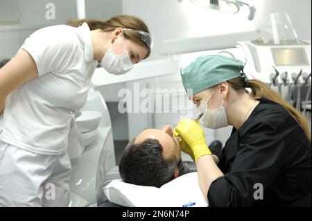 Le médecin de clinique dentaire enlève la dent patient dans des gants jaunes en noir vêtements assistant regarde la dernière technologie femme médecin et patient homme remplissage c Banque D'Images