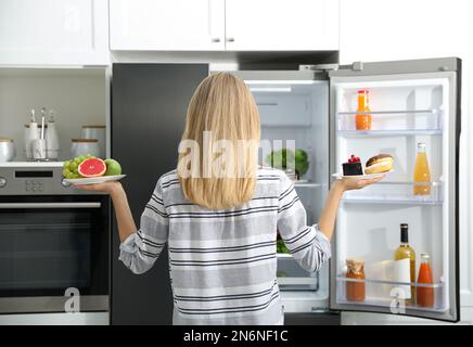 Concept de choix. Femme tenant des assiettes avec des fruits et des bonbons près du réfrigérateur dans la cuisine, vue arrière Banque D'Images