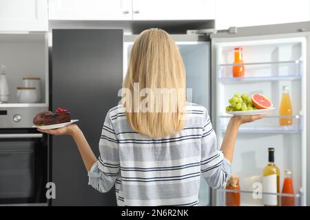 Concept de choix. Femme tenant des assiettes avec des fruits et des bonbons près du réfrigérateur dans la cuisine, vue arrière Banque D'Images