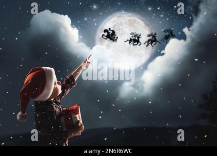 Joli petit garçon regardant le Père Noël avec des rennes dans le ciel la nuit pleine lune. Vacances de Noël Banque D'Images