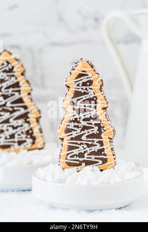 Deux biscuits en forme d'arbres de noël avec chocolat et sucre glacer dans des bols avec du sucre grossier sur fond blanc avec de la vaisselle blanche Banque D'Images