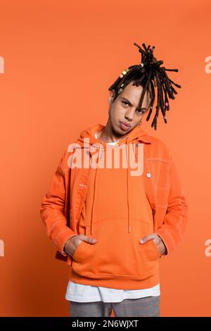 homme percé et multiracial avec des dreadlocks regardant l'appareil photo tout en se tenant avec les mains dans les poches sur fond de corail Banque D'Images