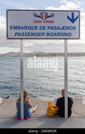 Deux touristes attendent le ferry, assis au pied du panneau indiquant la zone d'embarquement au port de Corralejo, Fuerteventura. Banque D'Images