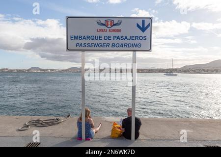 Deux touristes attendent le ferry, assis au pied du panneau indiquant la zone d'embarquement au port de Corralejo, Fuerteventura. Banque D'Images