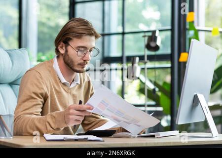 Un sérieux jeune homme comptable, auditeur, homme d'affaires est assis dans le bureau à la table. Concentrated fonctionne avec les documents, la calculatrice, les comptes, les plans. Banque D'Images