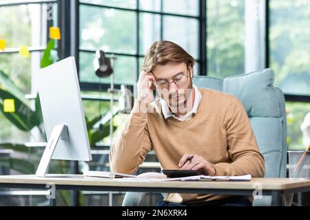 Un sérieux jeune homme comptable, auditeur, homme d'affaires est assis dans le bureau à la table. Concentrated fonctionne avec les documents, la calculatrice et les comptes. Pendu soigneusement sa tête sur sa main. Banque D'Images