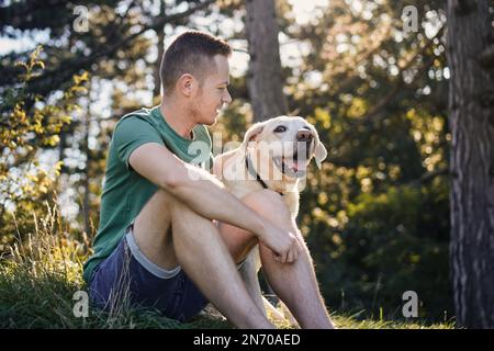 Homme avec son chien heureux assis dans l'herbe sous l'arbre. Propriétaire d'animal de compagnie appréciant le voyage avec son labrador retriever pendant la journée ensoleillée d'été. Banque D'Images