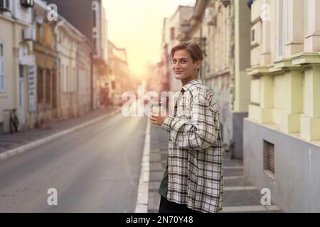 Une jeune femme urbaine se promenant dans la rue, tenant une tasse de café jetable recyclable dans la main, regardant l'appareil photo et souriant. Banque D'Images