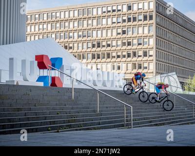 E-motards de montagne sur les marches de la Philharmonie avec le lettrage du Luxembourg. En arrière-plan, le bâtiment Schumann, construit en 70s pour abriter les premières institutions de l'UE à Luxembourg. Banque D'Images