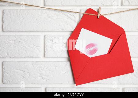 Enveloppe rouge et carte avec imprimé à lèvres accrochées à la ficelle près du mur de briques blanches, espace pour le texte. Lettre d'amour Banque D'Images