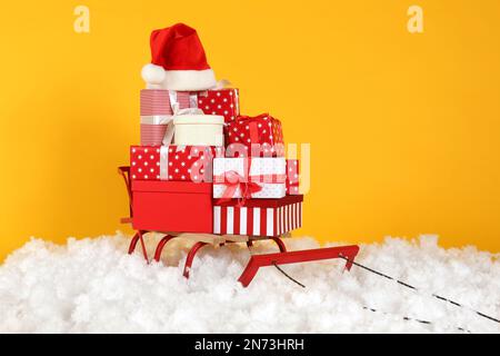 Traîneau avec cadeaux et chapeau de père Noël dans la neige artificielle sur fond jaune Banque D'Images