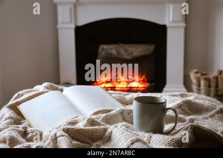 Tasse de thé chaud et livre près de la cheminée à la maison. Atmosphère chaleureuse Banque D'Images