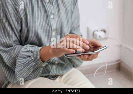 Femme utilisant un téléphone portable avec chargeur connecté près d'un mur de briques blanches à l'intérieur, gros plan Banque D'Images