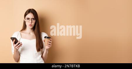 Une fille confuse dans des verres frognant à l'appareil photo après avoir lu un téléphone portable, tenant une tasse de café, debout déplaisée sur fond beige Banque D'Images