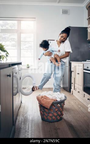 Une lessive pour nouveau-nés est abondante. une jeune mère utilisant un téléphone portable tout en effectuant des travaux ménagers et en tenant son bébé à la maison. Banque D'Images