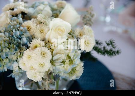 Roses blanches et hortensia bleu dans un vase en verre carré sur des nappes claires avec des napperons bleu marine, Banque D'Images