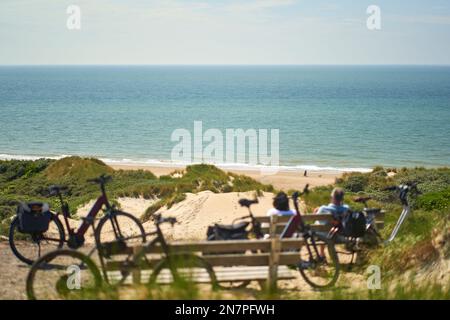 Plage de la mer du Nord en Hollande. Couple prenant une pause sur un banc en face des dunes de sable. Vélos électriques en avant-plan. Banque D'Images