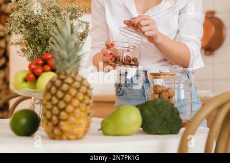 gros plan des mains de l'hôtesse tenant des noix dans un pot et des légumes couchés, des fruits au premier plan sur la table de cuisine. cuisson. Banque D'Images