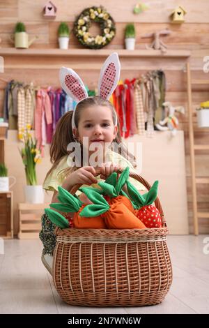 Adorable petite fille avec des oreilles de lapin et un panier rempli de carottes en peluche dans la zone de photo de Pâques Banque D'Images