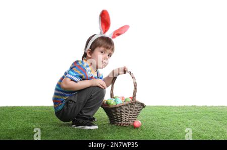 Un petit garçon mignon portant des oreilles de lapin avec un panier rempli d'œufs de Pâques teints sur de l'herbe verte Banque D'Images