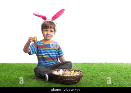 Un petit garçon mignon portant des oreilles de lapin avec un panier rempli d'œufs de Pâques teints sur de l'herbe verte Banque D'Images