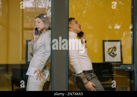 Deux collègues masculins et féminins attrayants ayant un appel téléphonique dans les cabines téléphoniques au bureau Banque D'Images