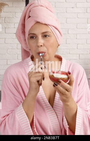une femme dans un peignoir après la douche mange un délicieux dessert pour le petit déjeuner à la maison. une femme d'âge moyen dans la cuisine apprécie son repas du matin. Banque D'Images