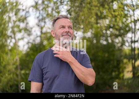 Homme pensif d'âge moyen debout devant des arbres verts avec sa main au cou ou à la gorge Banque D'Images