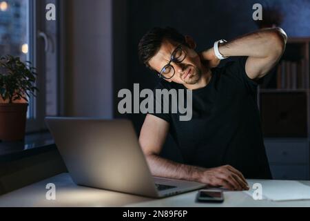 Un jeune homme d'affaires fatigué et stressé souffrant de douleurs à la nuque travaillant à domicile assis à une table avec un ordinateur portable la nuit. Un travailleur indépendant qui masse le cou se sentant mal à la douleur d'une posture incorrecte Banque D'Images