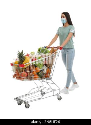 Jeune femme en masque médical avec chariot plein de provisions sur fond blanc Banque D'Images