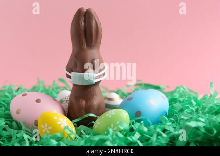 Lapin au chocolat avec masque de protection et œufs sur fond rose, espace pour le texte. Vacances de Pâques pendant la quarantaine COVID-19 Banque D'Images