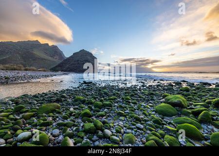 Le coucher du soleil depuis la plage rocheuse. La montagne sur la gauche, l'océan sur la droite, et la plage rocheuse avec des pierres vertes sur le fond. Personne. Quelques nuages. Banque D'Images