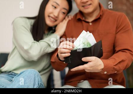 Concept d'économie d'argent. Heureux couple coréen mettant des billets dans un portefeuille, assis sur un canapé à la maison, attention sélective Banque D'Images