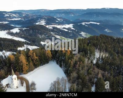 Une photo aérienne de l'église de Saint Primus et Felician au sommet d'une montagne forestière enneigée, Slovénie Banque D'Images