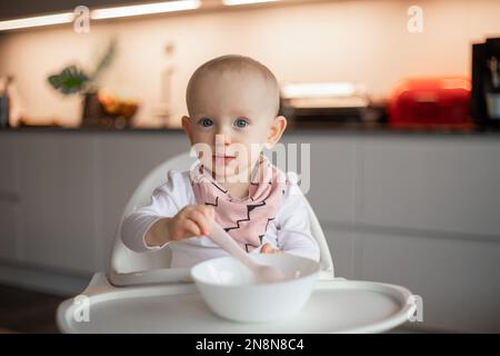 Un petit bébé heureux mange tout seul avec une cuillère tout en étant assis sur une chaise haute. Banque D'Images