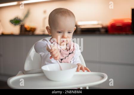 Un petit bébé heureux mange tout seul avec une cuillère tout en étant assis sur une chaise haute. Banque D'Images