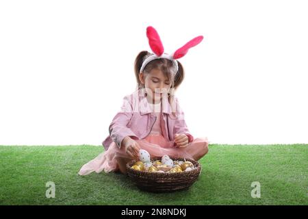 Adorable petite fille avec des oreilles de lapin et un panier en osier rempli d'oeufs de Pâques sur l'herbe verte sur fond blanc Banque D'Images