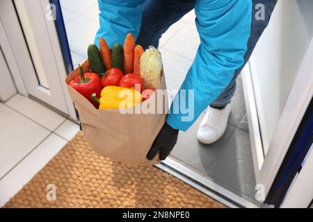 Transporteur tenant le sac en papier avec la nourriture dans la porte, gros plan. Service de livraison pendant la quarantaine en raison de l'éclosion de Covid-19 Banque D'Images