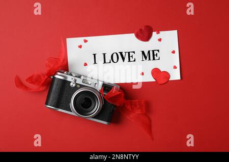 Papier avec la phrase manuscrite I Love Me, appareil photo et coeurs sur fond rouge, couché plat Banque D'Images