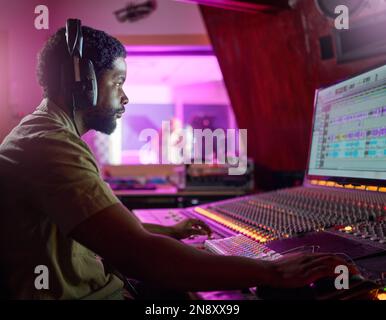 Homme noir, enregistrement en studio et travail musical la nuit avec casque, ordinateur ou Focus en mélangeant la console. Ingénieur, producteur ou technologie audio Banque D'Images