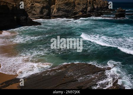 Surf blanc sur des vagues vertes de l'Atlantique qui survolent une plage de sable et qui coulent sur une étagère de roches sédimentaires jurassiques tôt dans une crique déserte près de la pointe de Cabo Carvoeiro, Peniche, district de Leiria, Portugal. Banque D'Images