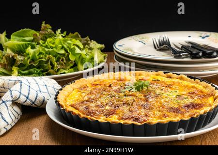 Vue latérale de quiche de bacon et d'épinards maison dans une tarte métallique noire au four plaque de cuisson sur un plat en céramique blanc avec légumes verts, nappe, pile de Banque D'Images