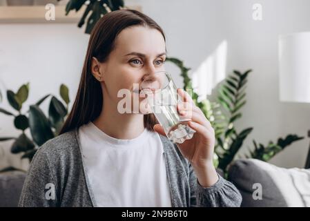 Souriant belle et paisible jeune femme enceinte boire un verre d'eau minérale fraîche et pure assis sur le canapé à la maison, empêchant la déshydratation de profiter Banque D'Images