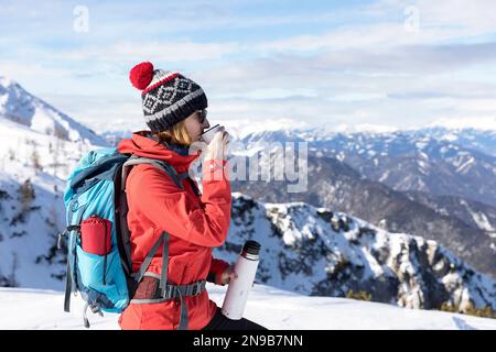 Une randonneuse touristique boit du thé chaud lors d'une randonnée hivernale sur le mont Dobratsch, profitant de vues panoramiques spectaculaires enneigées en Autriche. Banque D'Images